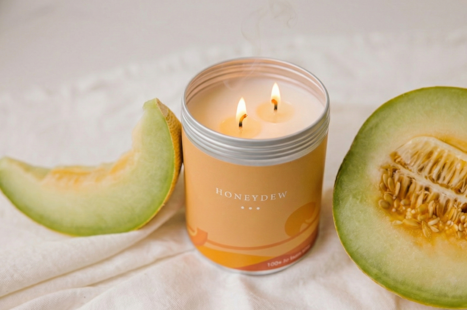 Honeydew candle in a tin with two lit candles, surrounded by honeydew melon slices on a white background.