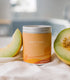 A Large Orange Tin With Melon Designs on the side. Made in Kalamazoo, MI. Scented with Melons, Honey, and Earth.