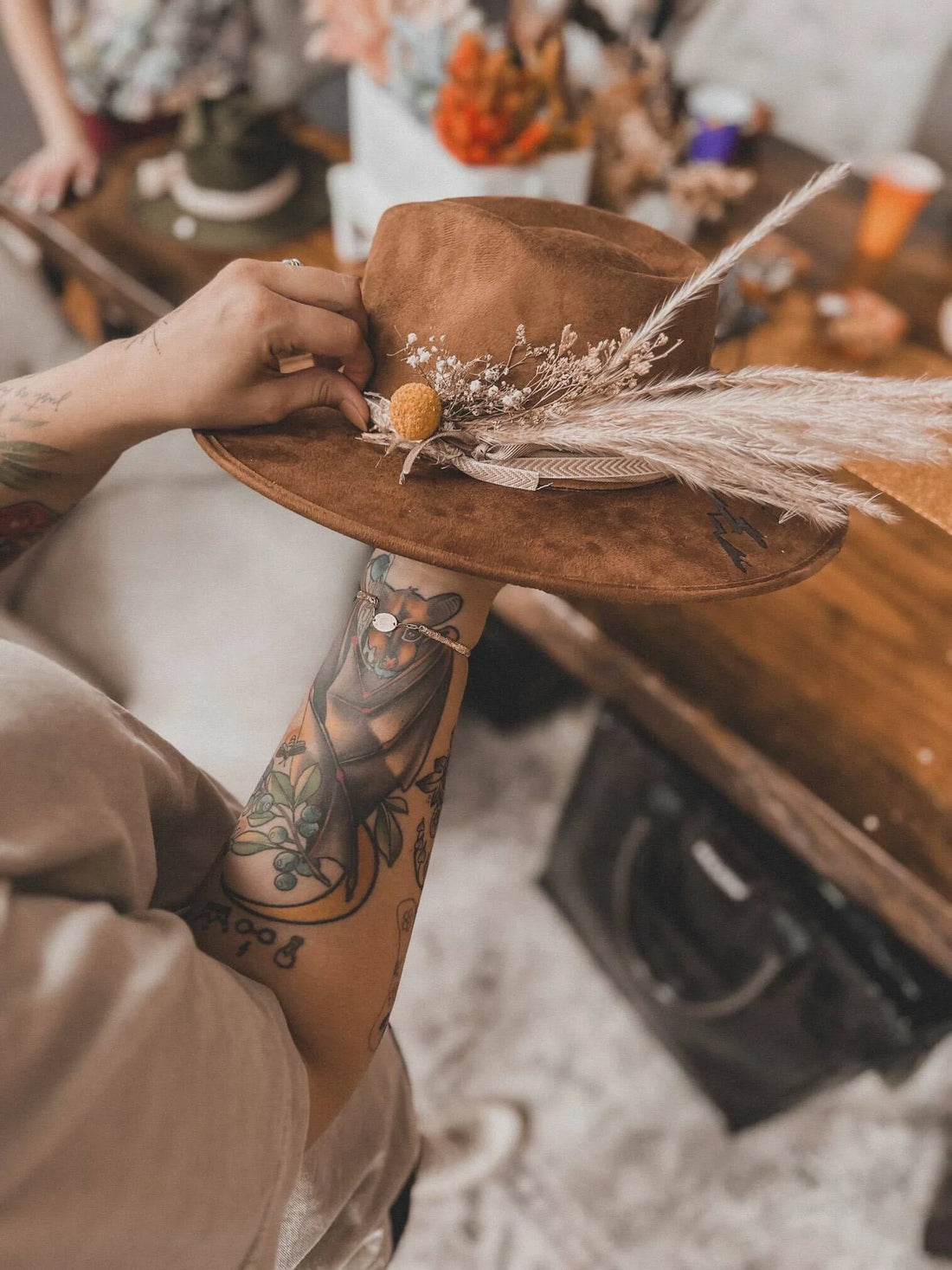 close up photo of a suede wide brimmed hat with feathers, dried flowers held by someone with a forearm tattoo