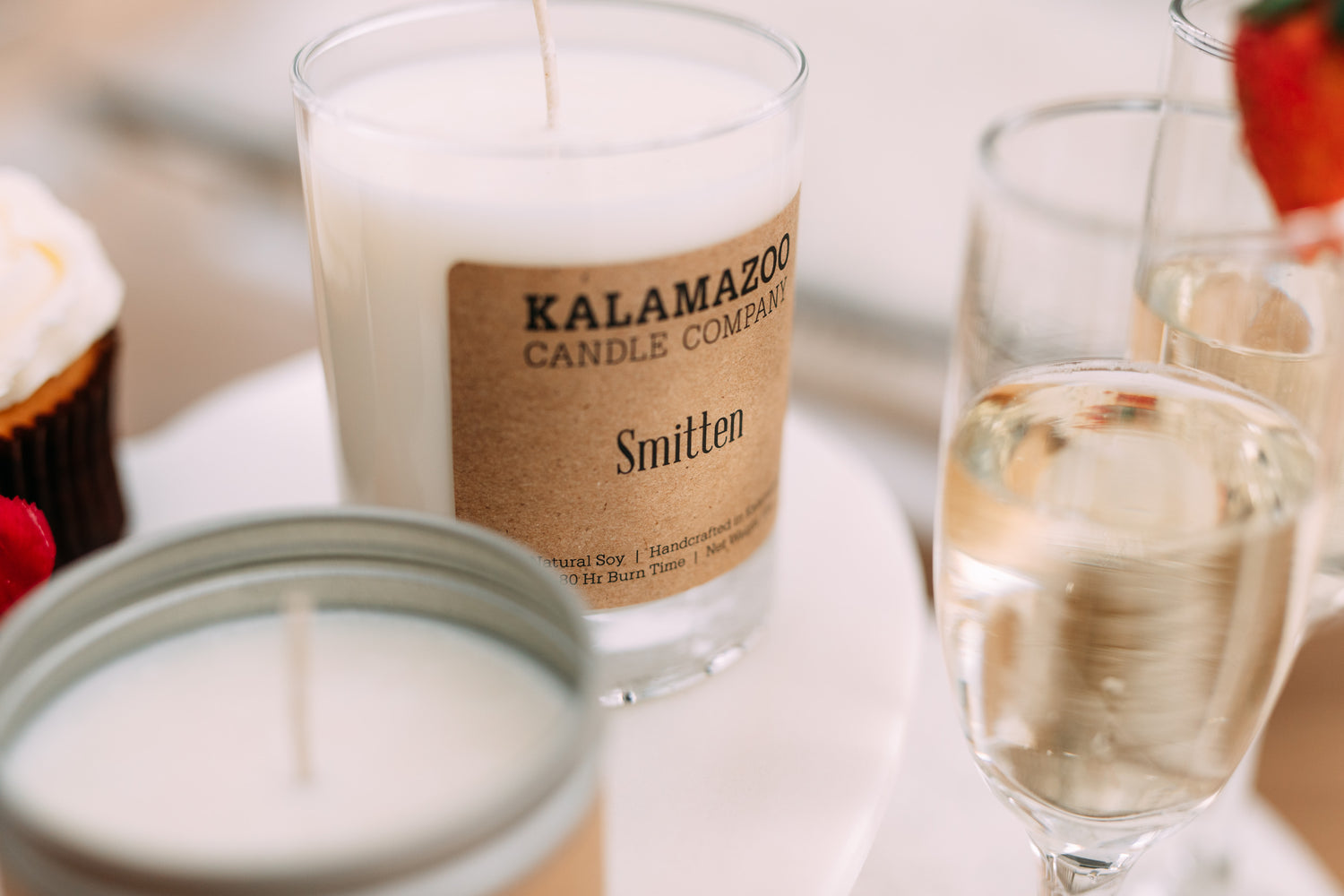 clear jar with off white candle wax with a craft paper label with the words Kalamazoo Candle Company in the scent "Smitten. Candle is surrounded by blurred images of champagne glasses filled halfway, vanilla cupcake with vanilla frosting. 