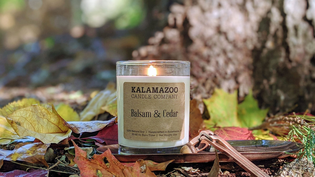 Lit Balsam & Cedar soy candle among colorful fall leaves beside a birch tree