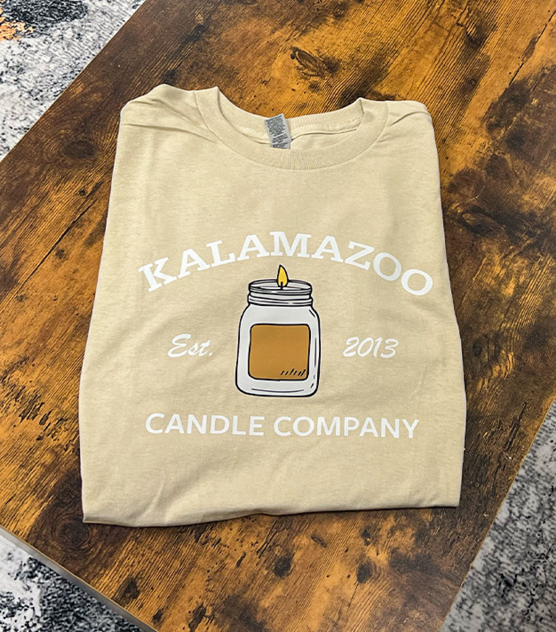 A tan Kalamazoo Candle T-Shirt with a candle on the front.
